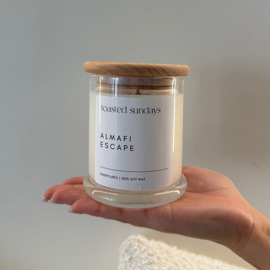Hand holding a candle labeled 'Almalfi Escape' by 'Toasted Sundays' against a plain background