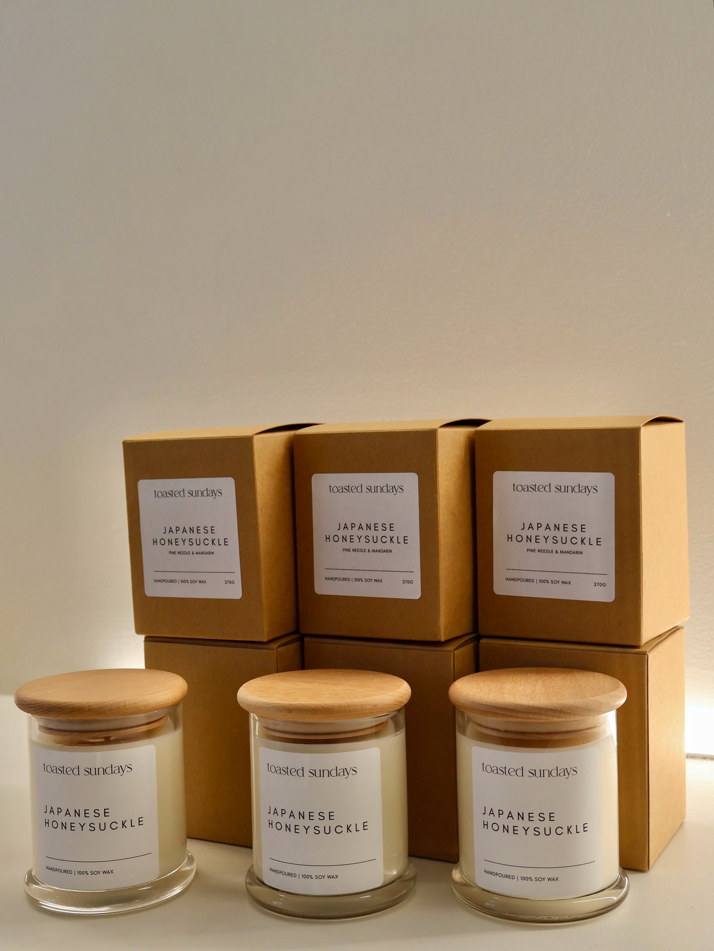 Three candles with wooden lids and boxes labeled 'Japanese Honeysuckle' on a neutral background.