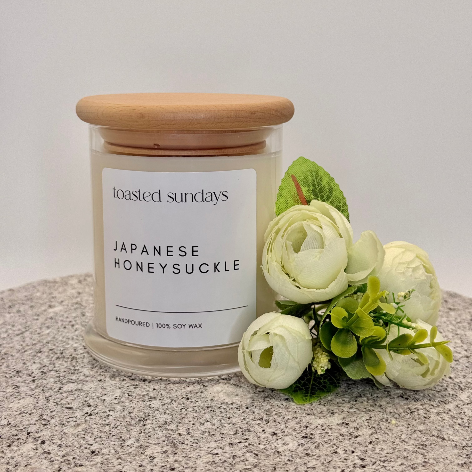 Candle labeled 'Toasted Sundays Japanese Honeysuckle' with a wooden lid on a surface with flowers.