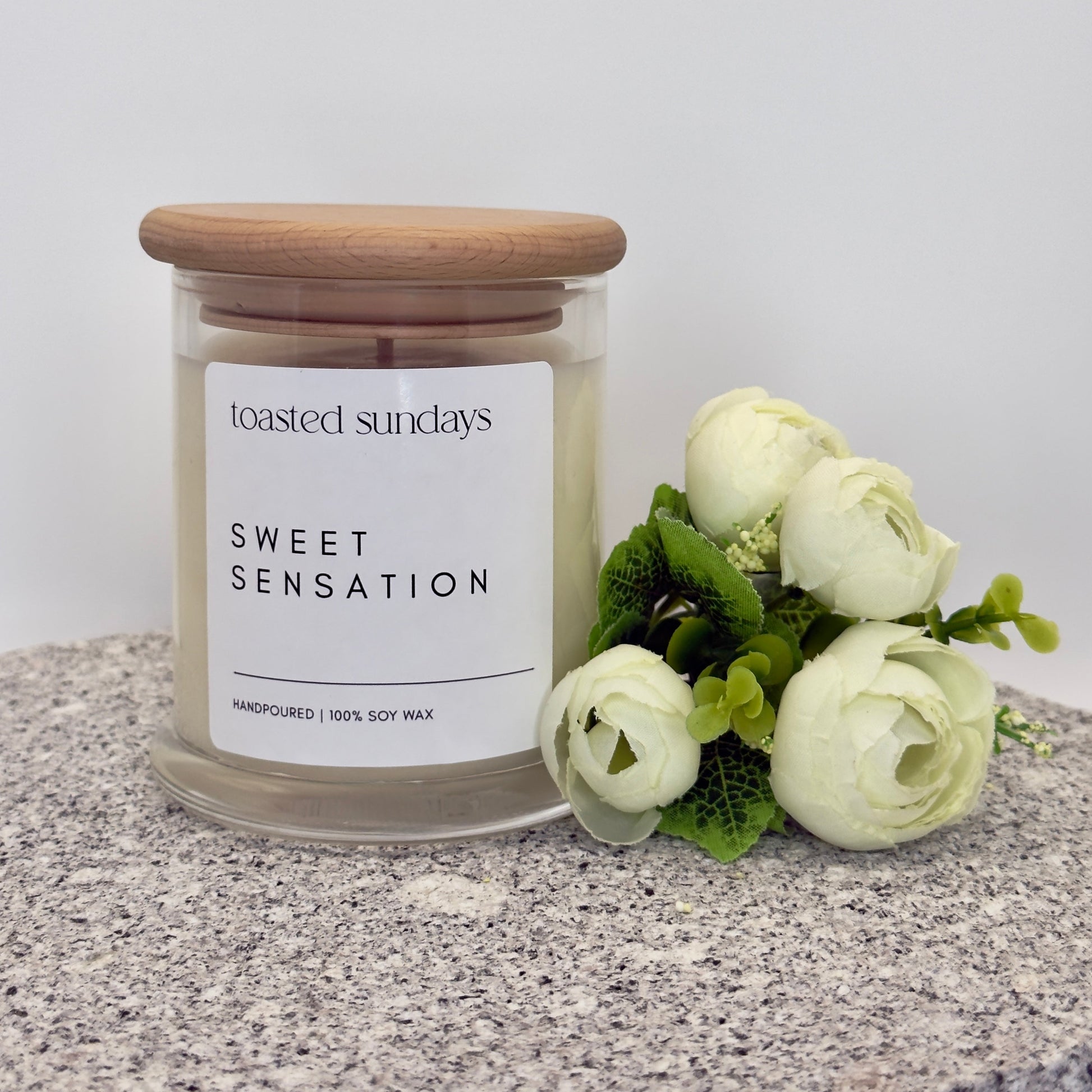 Candle labeled 'Toasted Sundays Sweet Sensation' with a wooden lid on a textured surface with flowers.
