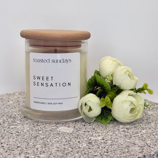 Candle labeled 'Toasted Sundays Sweet Sensation' with a wooden lid on a textured surface with flowers.