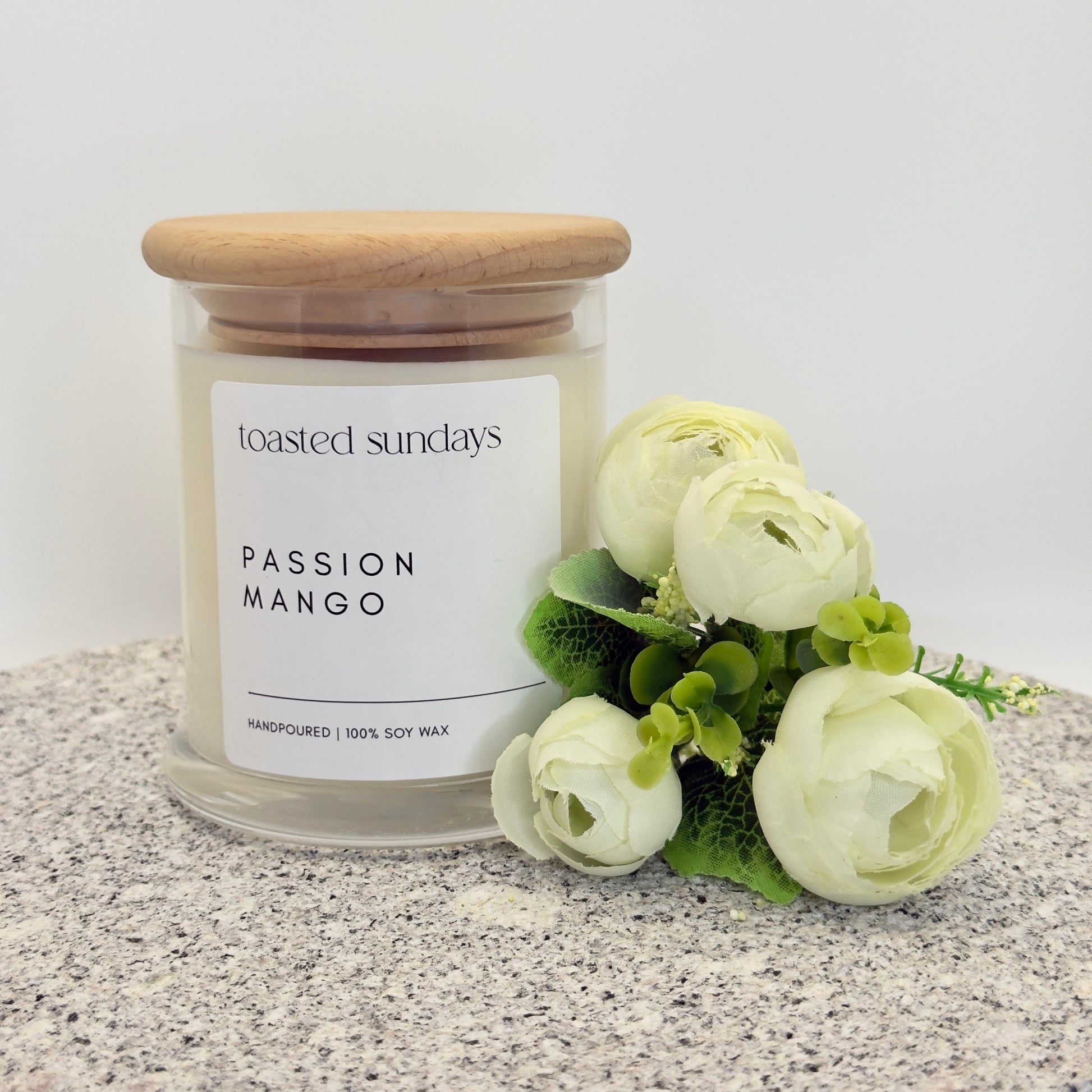 Candle labeled 'Toasted Sundays Passion Mango' with a wooden lid on a textured surface with flowers.