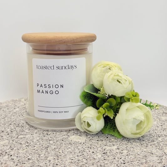 Candle labeled 'Toasted Sundays Passion Mango' with a wooden lid on a textured surface with flowers.