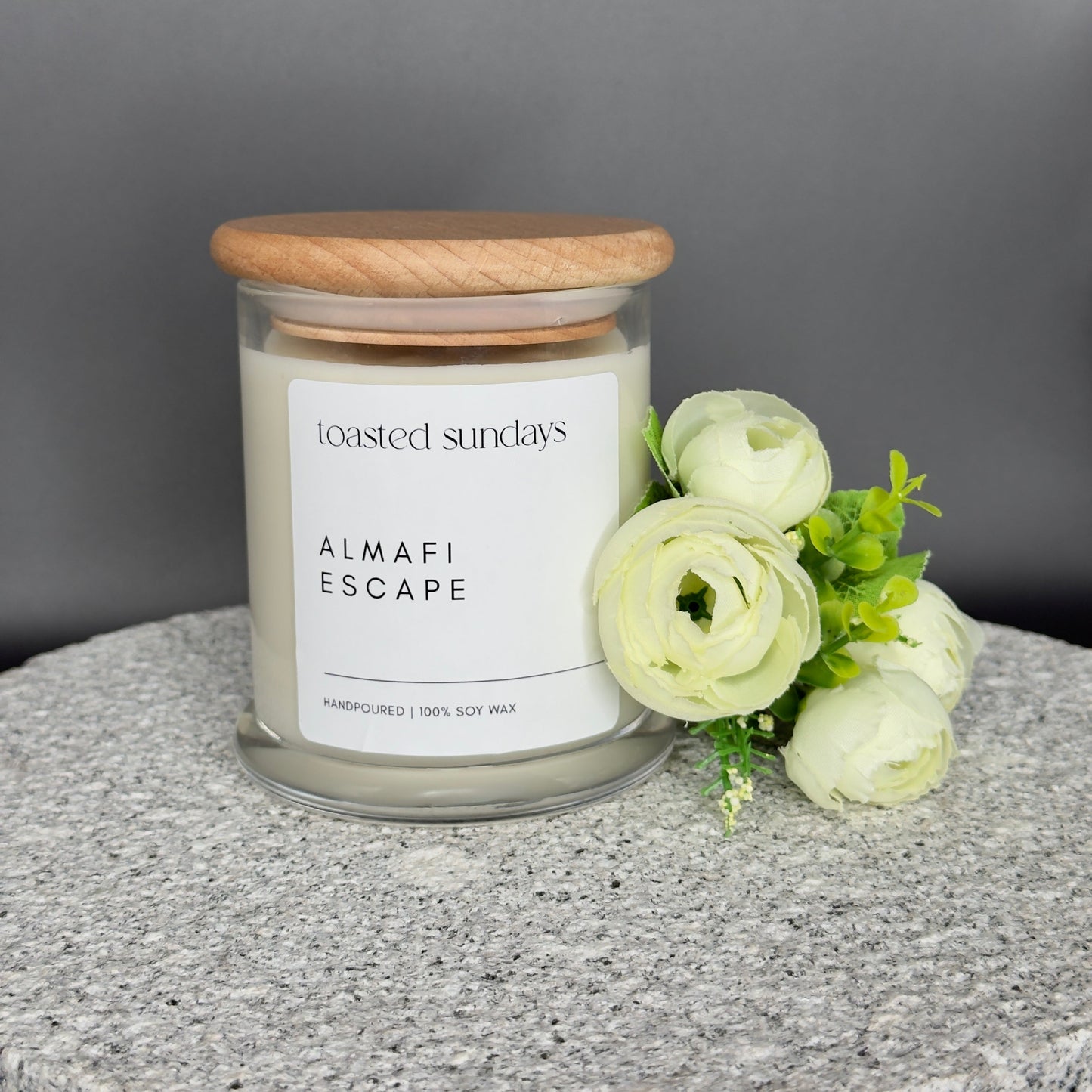 Candle jar labeled 'Toasted Sundays Almami Escape' with a wooden lid on a gray surface, accompanied by white flowers.