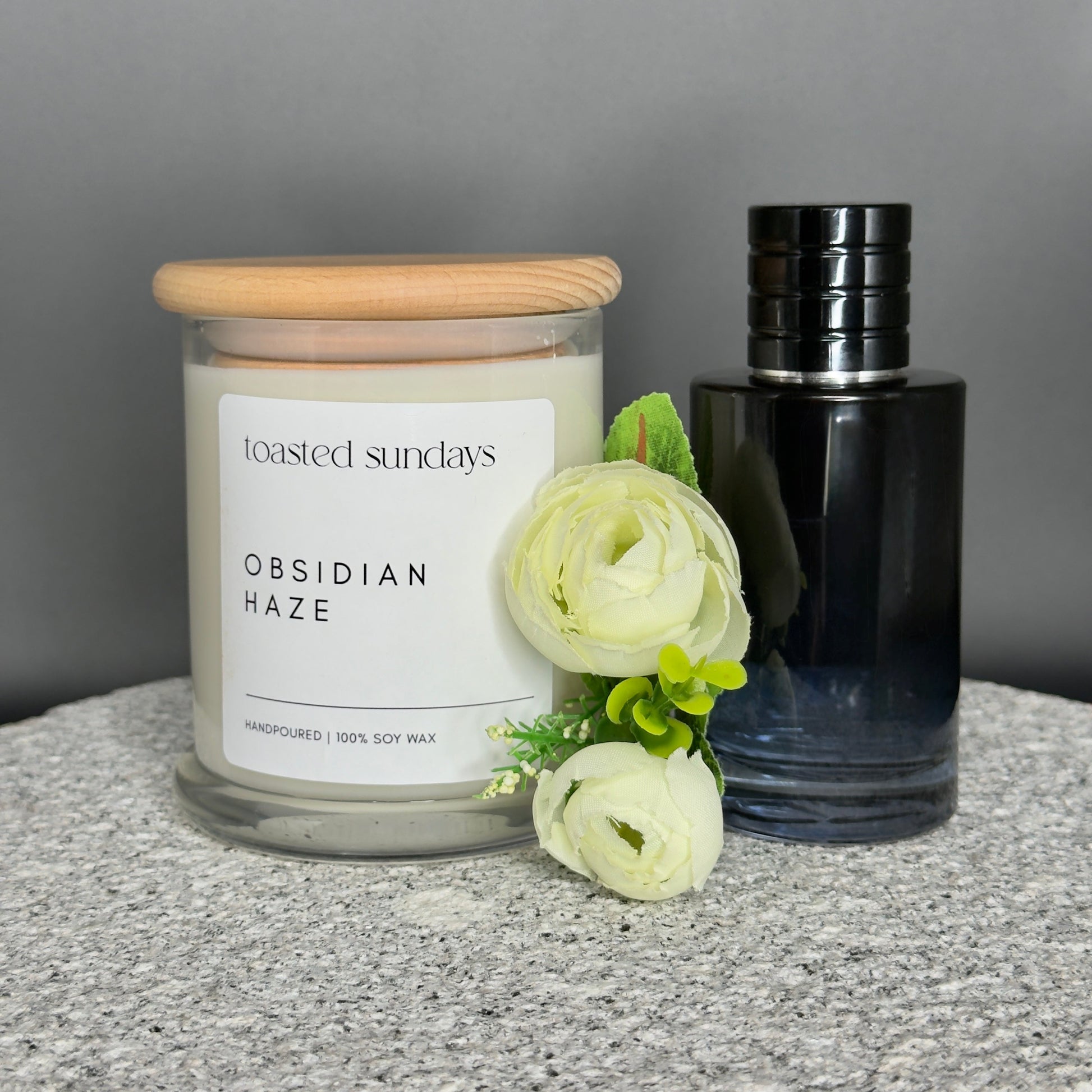 Candle labeled 'Obsidian Haze' by 'Toasted Sundays' with a black bottle and white flowers on a gray surface.