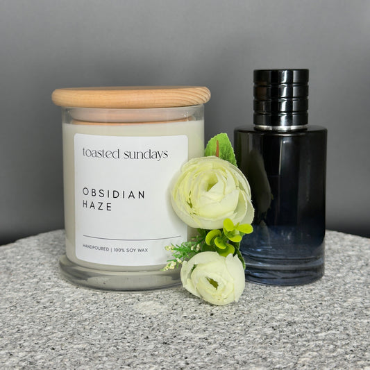 Candle labeled 'Obsidian Haze' by 'Toasted Sundays' with a black bottle and white flowers on a gray surface.