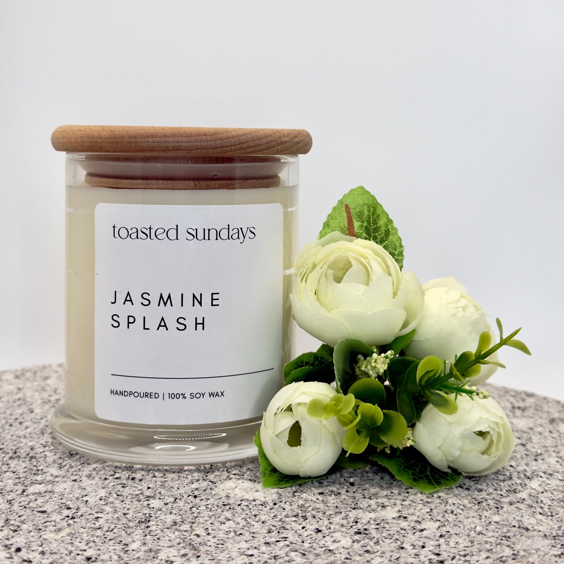 Candle labeled 'Toasted Sundays Jasmine Splash' with a wooden lid and white flowers on a gray surface.