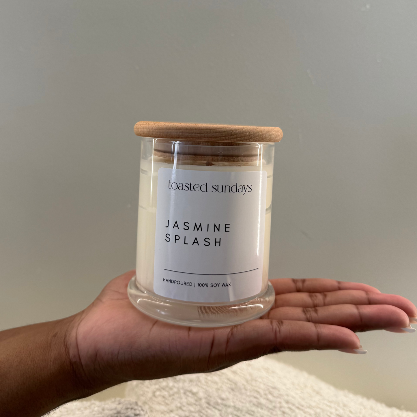 Hand holding a candle labelled 'Jasmine Splash' by 'Toasted Sundays' against a neutral background.