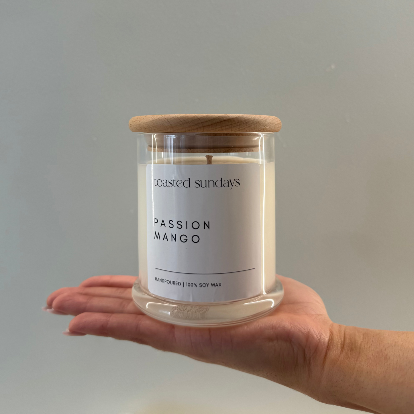 Hand holding a 'Passion Mango' candle with a wooden lid against a plain background