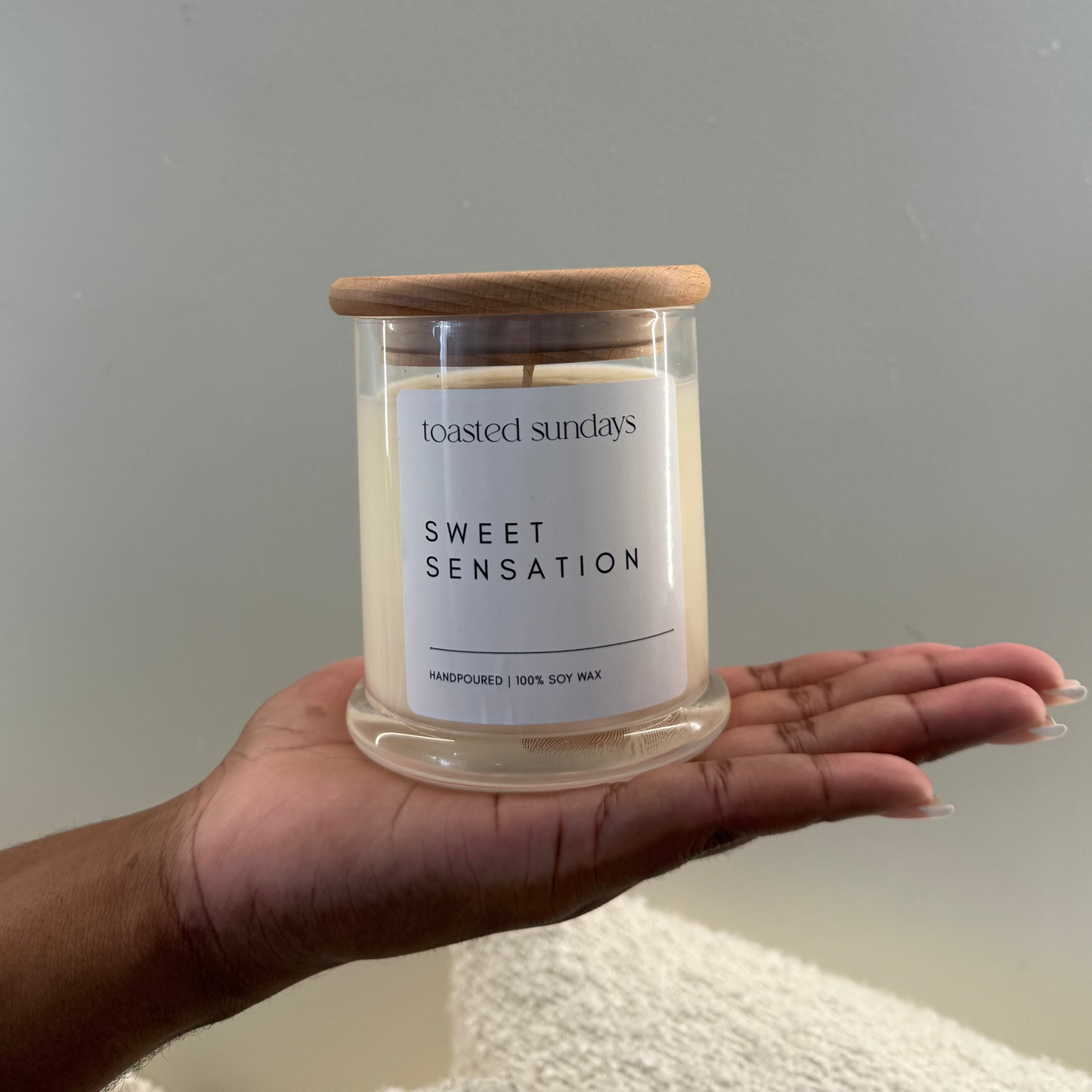 Hand holding a 'Sweet Sensation' candle with a wooden lid against a plain background